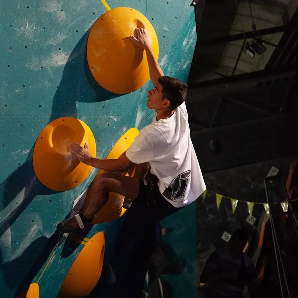Een jonge man beklimt een bouldermuur in een klimhal; hij houdt zich met beide handen en één voet vast aan grote gele grepen en draagt een wit T-shirt en een donkere korte broek. De muur is blauwgroen en de klimhal is schemerig verlicht. Een jonge man beklimt een bouldermuur in een klimhal; hij houdt zich met beide handen en één voet vast aan grote gele grepen en draagt een wit T-shirt en een donkere korte broek. De muur is blauwgroen en de klimhal is schemerig verlicht.
