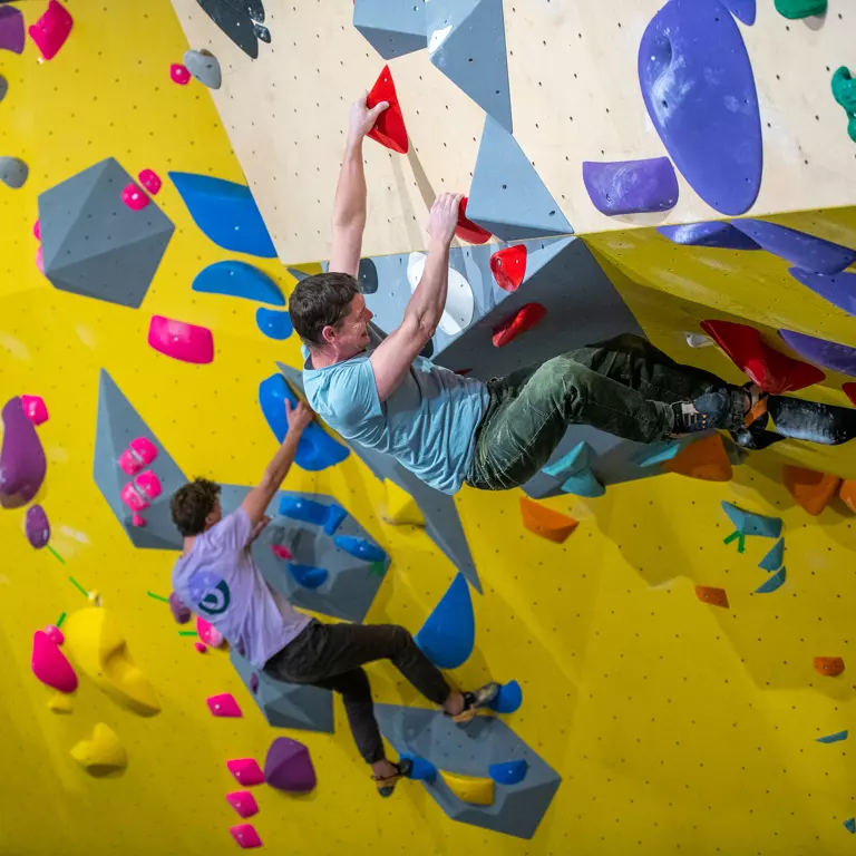 Twee mensen zijn aan het klimmen op een felgele bouldermuur met kleurrijke hand- en voetsteunen. Ze zijn allebei bezig met klimmen, houden zich vast aan verschillende grepen en zien er geconcentreerd uit. Twee mensen zijn aan het klimmen op een felgele bouldermuur met kleurrijke hand- en voetsteunen. Ze zijn allebei bezig met klimmen, houden zich vast aan verschillende grepen en zien er geconcentreerd uit.