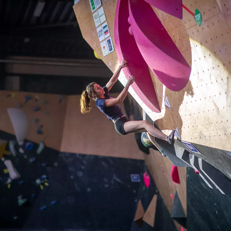 Een vrouw met rood haar beklimt een indoor-bouldermuur en houdt zich vast aan grote roze grepen. Ze ziet er geconcentreerd en atletisch uit, met één voet op een kleine voetsteun en wedstrijdstickers die zichtbaar zijn op de klimwand. Een vrouw met rood haar beklimt een indoor-bouldermuur en houdt zich vast aan grote roze grepen. Ze ziet er geconcentreerd en atletisch uit, met één voet op een kleine voetsteun en wedstrijdstickers die zichtbaar zijn op de klimwand.