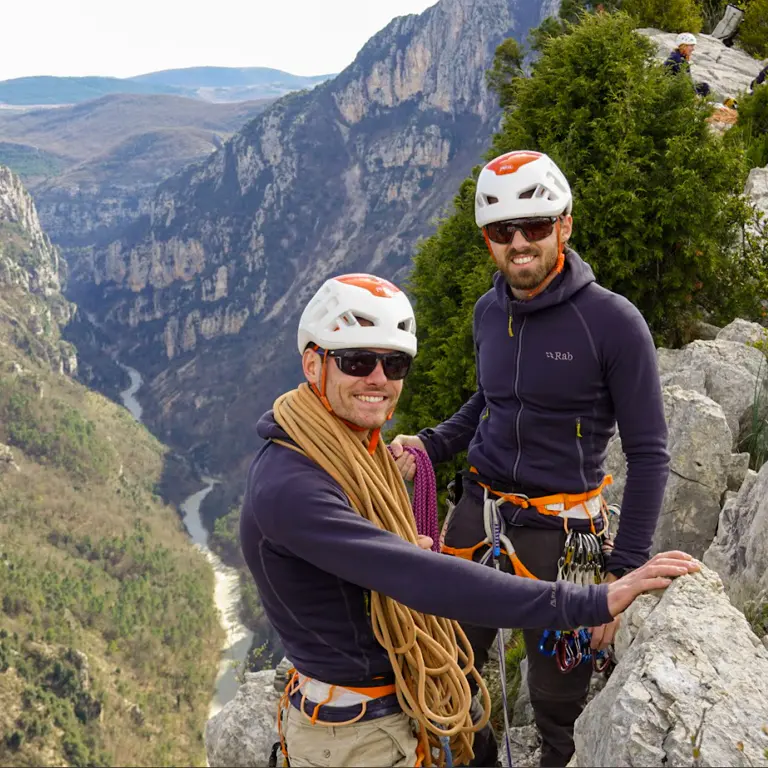 Twee bergbeklimmers met helmen en klimgordels poseren op de rand van een klif die uitkijkt over een rivierkloof. Twee bergbeklimmers met helmen en klimgordels poseren op de rand van een klif die uitkijkt over een rivierkloof.